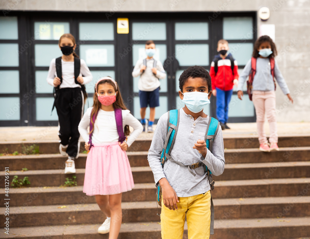 Obraz premium Portrait of schoolboy in medical mask standing near school, kids on background ..