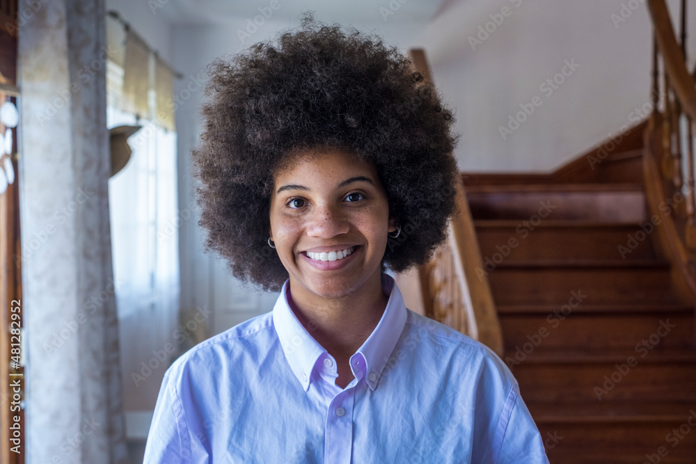 Portrait of smiling millennial attractive african american woman at home. Pretty happy black lady looking at camera in domestic room of house. Pleased female with curly hairstyle in modern apartment
