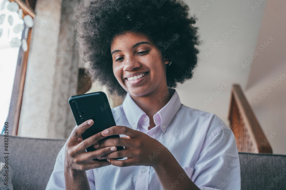 Smiling millennial african american woman using mobile phone at home. Happy black lady with curly hairstyle text messaging or chatting on smartphone. Young female browsing social media content 