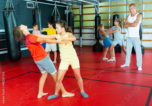 Kids exercising self-protection moves together on their training. Their teacher standing nearby and observing.
