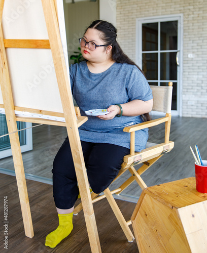 Asian young cute fun chubby down syndrome autistic female art artist student wearing eyeglasses sitting smiling at table holding color pencil drawing picture on paper in front brick wall background