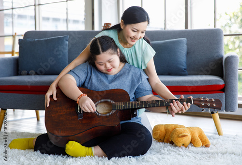 Foto Asian lovely mother helping young chubby down syndrome autistic autism little daughter holding playing guitar in living room at home