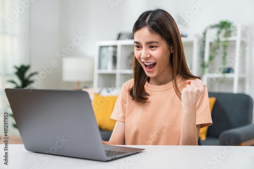 Cheerful beautiful Asian woman using laptop with fists clenched celebrating victory expressing success.