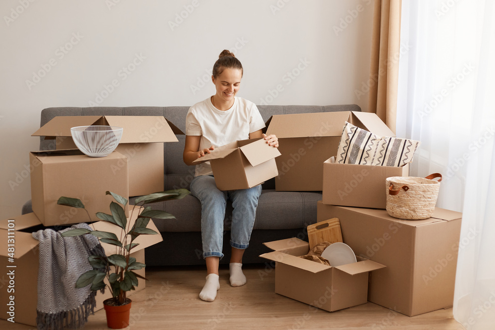 Indoor shot of positive smiling woman wearing white t-shirt and jeans sitting on sofa in a new house or flat after relocating, holding cardboard box, looking inside, unpacking packages. Indoor shot of positive smiling woman wearing white t-shirt and jeans sitting on sofa in a new house or flat after relocating, holding cardboard box, looking inside, unpacking packages.