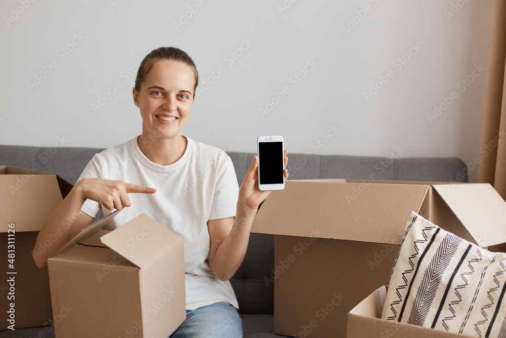 Smiling woman wearing white t-shirt and jeans sitting on sofa, posing in a new apartment after relocation, pointing at smartphone with empty screen for advertisement, removals and delivery service. Smiling woman wearing white t-shirt and jeans sitting on sofa, posing in a new apartment after relocation, pointing at smartphone with empty screen for advertisement, removals and delivery service.