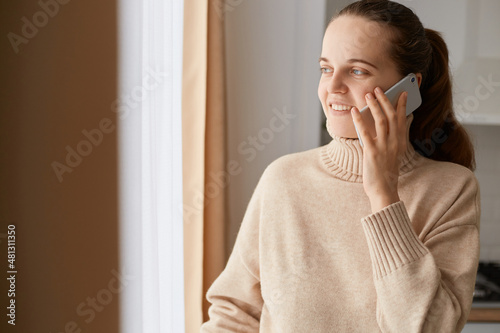 Photos Indoor shot of adorable smiling woman with ponytail hairstyle wearing beige casual style sweater talking phone with positive facial expression, female gossiping with friend via cell phone