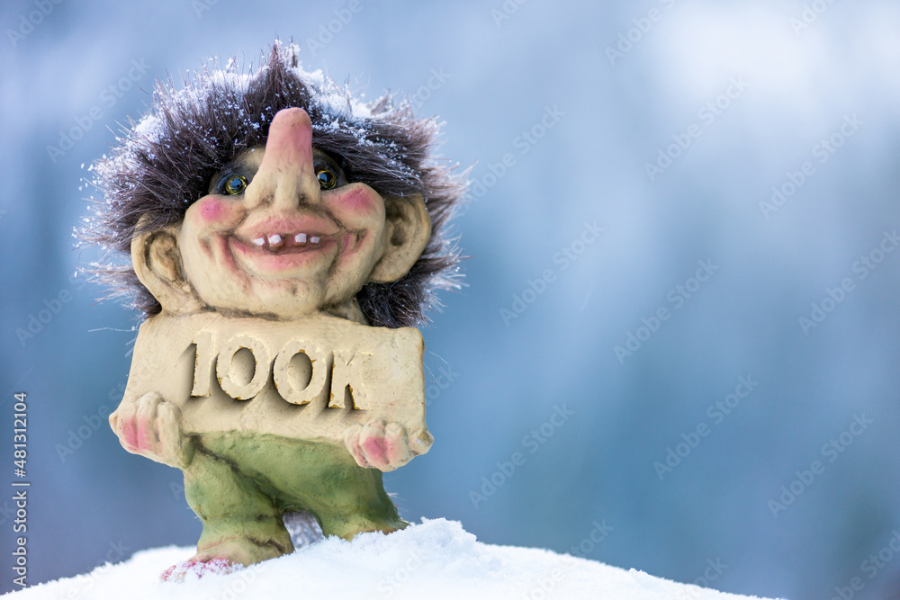Cute happy troll holding sign with the word 100k chiseled out. Soft blurred out background in winter wonderland. Cute happy troll holding sign with the word 100k chiseled out. Soft blurred out background in winter wonderland.