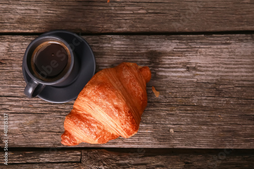 Expresso cup and croissant on old rustic wooden table background. Fresh baked bun and hot black coffee top view. Food, French breakfast, good morning, cafe menu concept. Close up, copy space