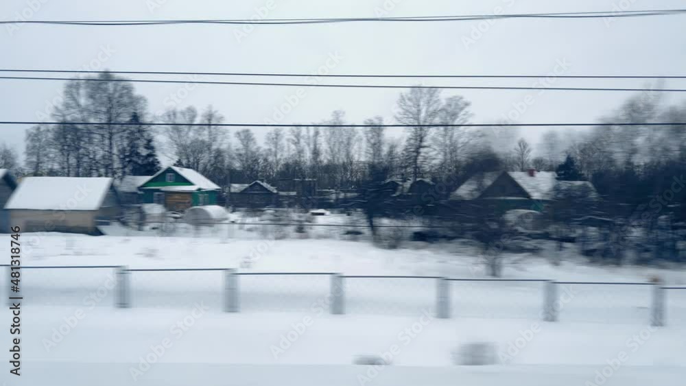 View from the window of moving train on snow-covered village one-storey houses, fence, trees on cloudy cold day