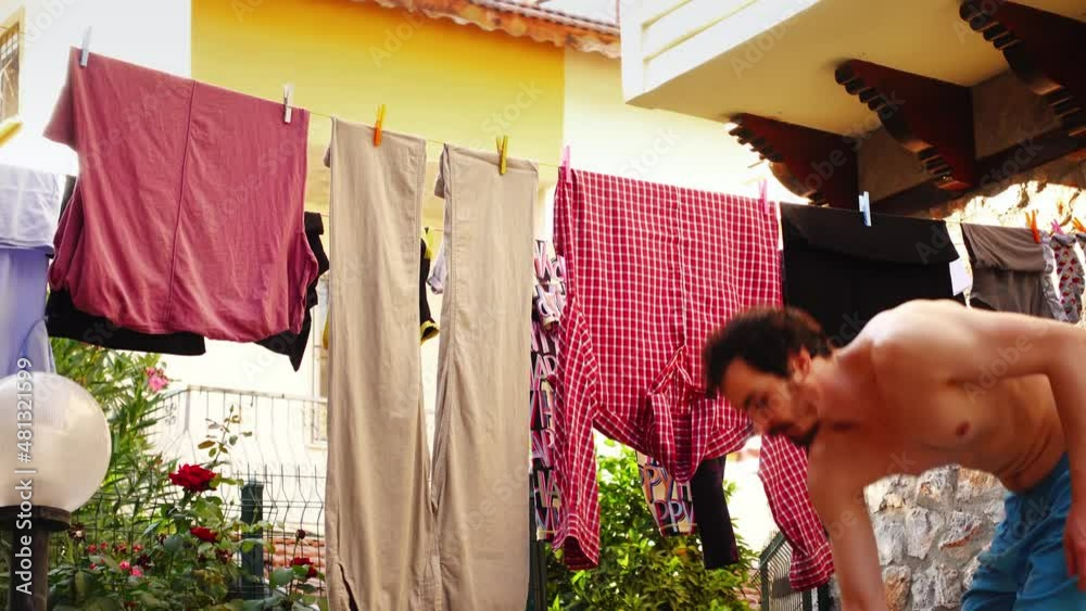 Young topless man in shorts is taking off clean dry clothes after laundry from the line and putting them tidily to basket. A caucasian bearded person washed his textile and got fresh pants and shirts.