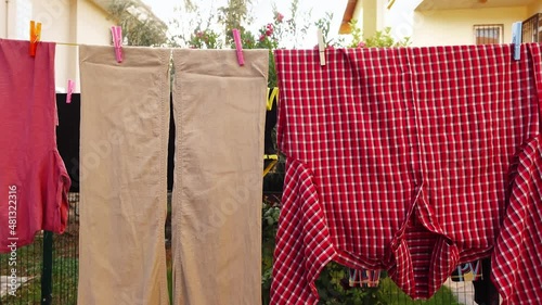 Beige pants and red checkered shirt are hanging on the clothesline attached by pins after laundry outdoors. Linen textile is drying and trembled by wind after being washed on the backyard of the house photo