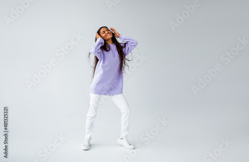 Beautiful brunette girl with curly hairstyle in purple hoodie and white sneakers and jeans posing on light background. black woman listening to music with headphones