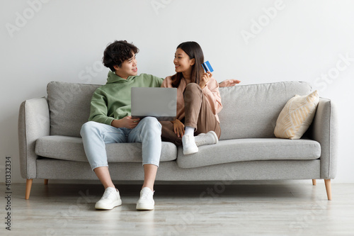 Happy young Asian spouses shopping online via laptop, using credit card, sitting on sofa at home, empty space