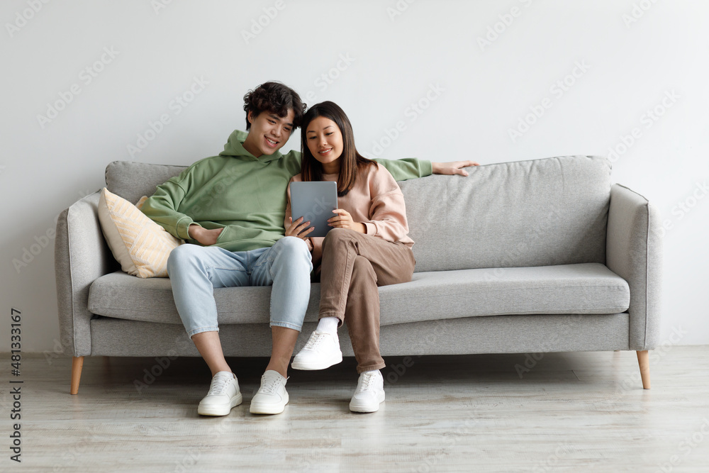 Happy Asian couple sitting on couch with tablet, looking at touch pad screen, using newest application at home Happy Asian couple sitting on couch with tablet, looking at touch pad screen, using newest application at home