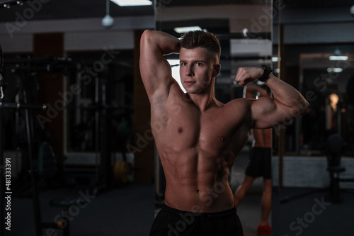 strong young man bodybuilder with a hairstyle and a bare muscular torso trains and poses in the gym against a dark background