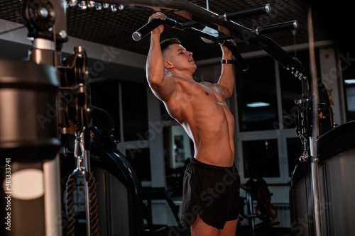 Handsome sporty guy trainer with tanned muscular naked torso pulls up in the gym