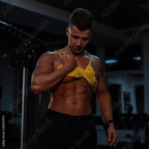 Sexy muscular guy athlete with a tanned body lifted a T-shirt and shows abdominal muscles on a dark background. Bodybuilder training in the gym