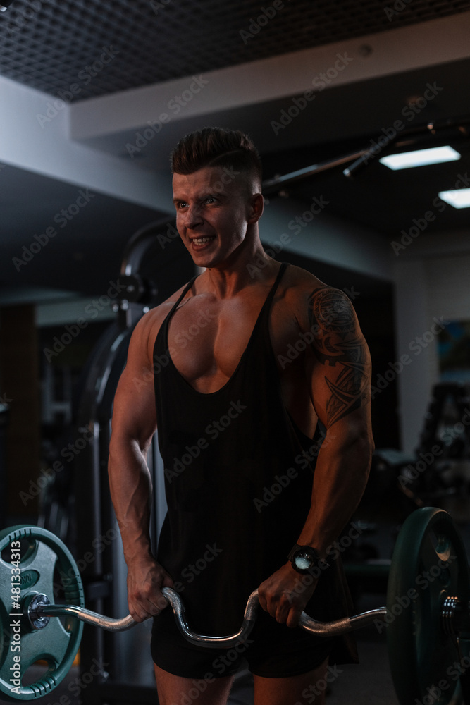 Strong athletic bodybuilder man in a black t-shirt pumps muscles in the gym on a dark background Strong athletic bodybuilder man in a black t-shirt pumps muscles in the gym on a dark background