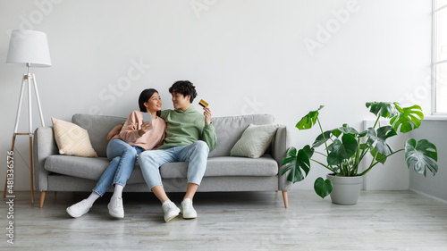 Cheerful young Asian couple sitting on sofa with credit card and cellphone, shopping online in living room, copy space