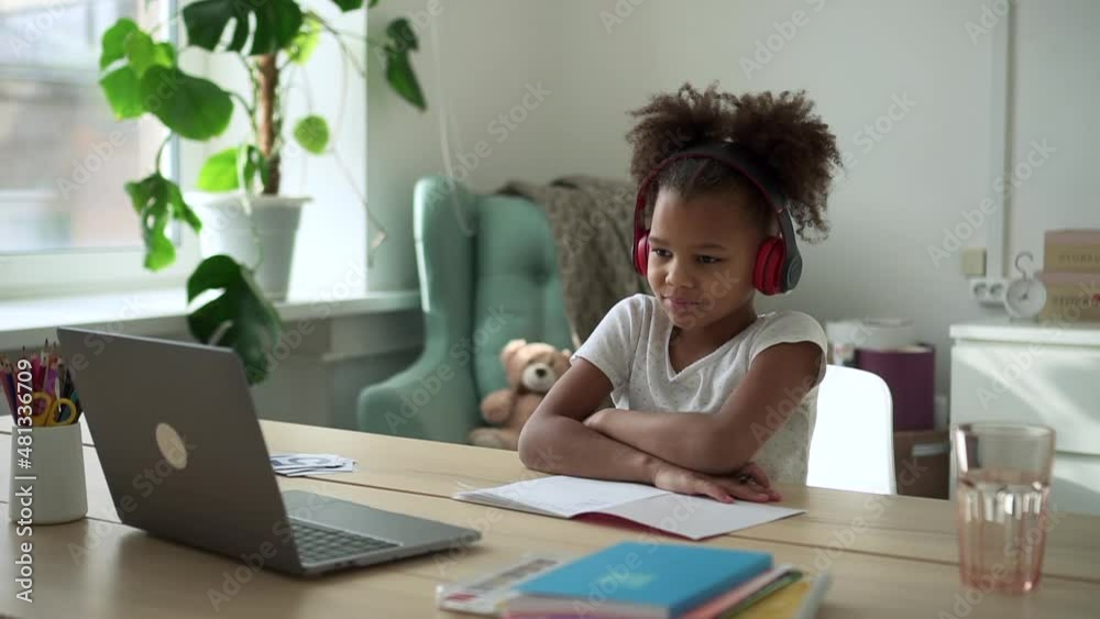 Little schoolgirl studying in online class and sitting at table with laptop in home room spbd. Close-up view of American African girl watches and listens to video lecture on computer screen, raises Little schoolgirl studying in online class and sitting at table with laptop in home room spbd. Close-up view of American African girl watches and listens to video lecture on computer screen, raises