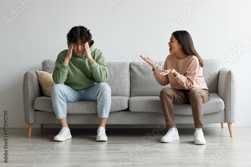 Angry millennial Asian wife yelling at her husband, man suffering from stress, sitting on sofa in living room, free space