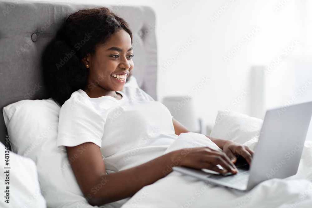 Smiling young black woman typing on laptop, working in bed Smiling young black woman typing on laptop, working in bed