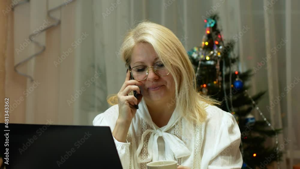 Smiling Caucasian blonde over 40 years old talking on the phone, doing remote work while sitting near a laptop against the backdrop of a New Year's festive interior at home. Freelancer with a phone Smiling Caucasian blonde over 40 years old talking on the phone, doing remote work while sitting near a laptop against the backdrop of a New Year's festive interior at home. Freelancer with a phone
