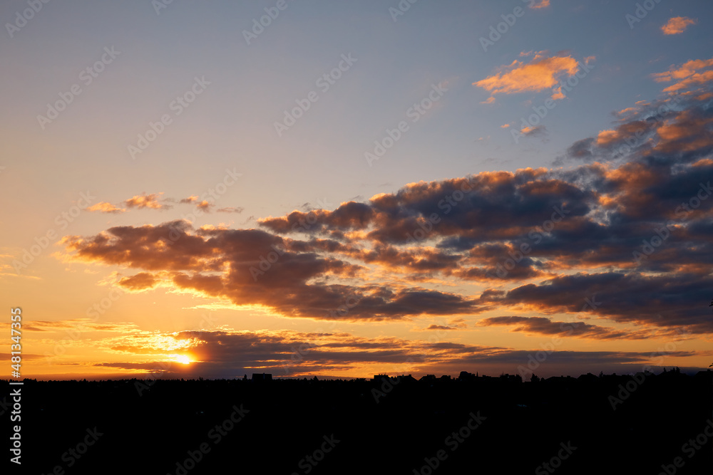 Landscape of the evening sunset sky with the sun in the clouds over a small town or village. Orange and blue sunrise. Beautiful scenery wallpaper. Early morning. Sunny weather. Silhouette of horizon