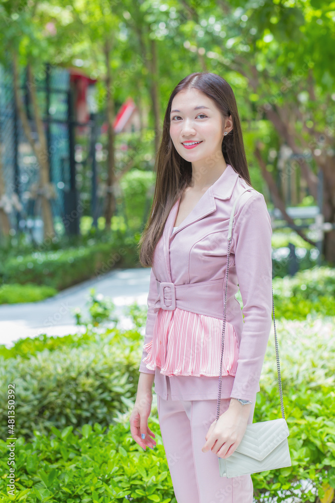 Beautiful young Asian professional business woman with long hair wearing pink shirt is smiling outdoor in the garden front of the office with trees background. Beautiful young Asian professional business woman with long hair wearing pink shirt is smiling outdoor in the garden front of the office with trees background.