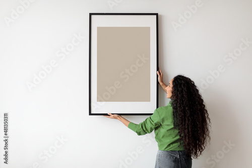 Foto Happy Woman Hanging Empty Poster In Frame On Wall Indoors