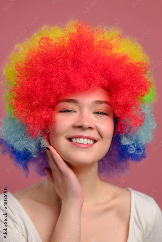 Portrait of woman with rainbow curly hairstyle. Smiling young attractive female in colorful wig on pink background. Close-up.