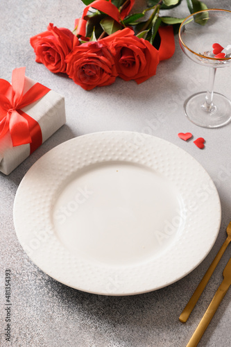 Valentine's day place setting with red roses and romantic gift on gray table. Vertical orientation. Festive romantic dinner.