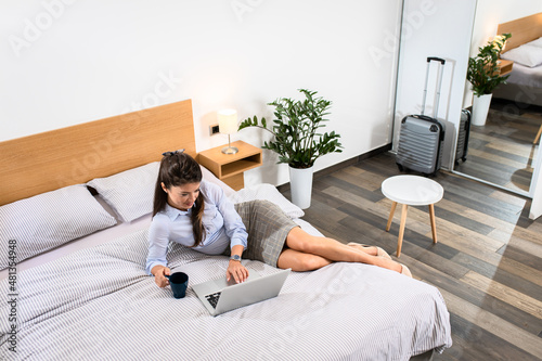 Businesswoman in hotel room on business trip sitting on bed using laptop.