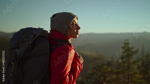 Side portrait woman hiker with backpack standing in sunset light among mountains and hills, close up slow motion. successful ascent on top of mountain. photo