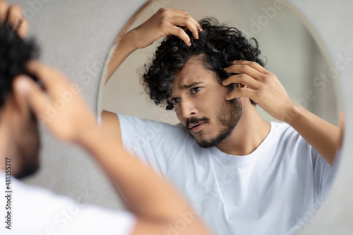Haircare treatment. Worried indian guy looking at flaky scalp in mirror, examining gray hair and hairloss issue photo