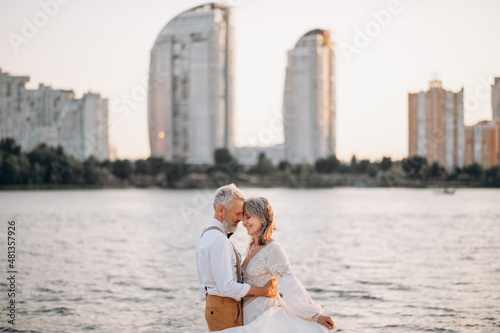 Senior newlyweds hug. Stylish couple of elderly newlyweds stand embracing on river bank. © MZaitsev