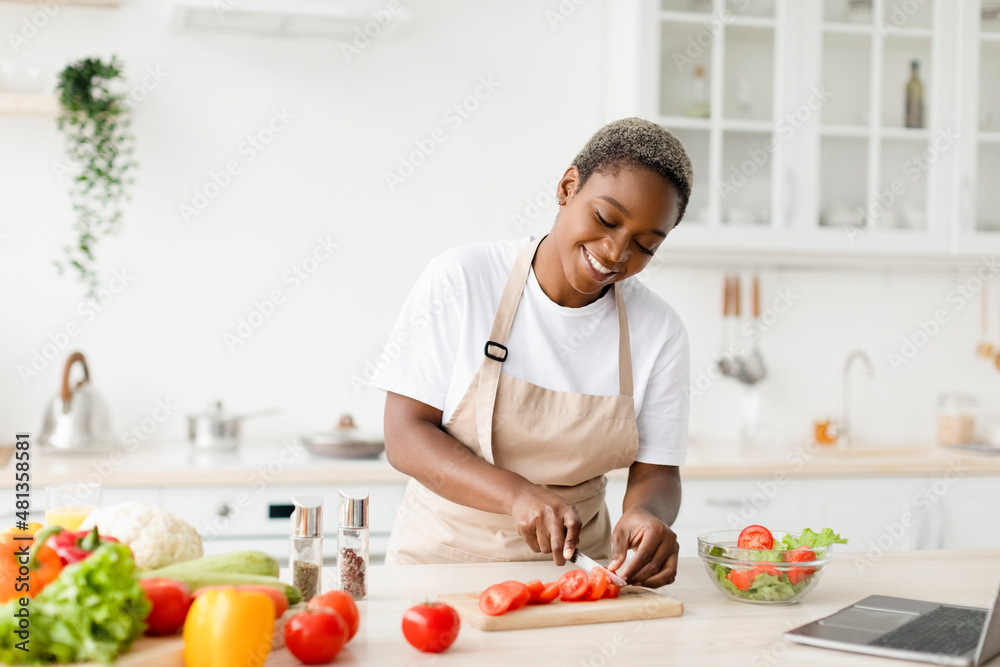 Smiling millennial pretty black female in apron prepare healthy lunch from organic vegetables, cuts tomatoes Smiling millennial pretty black female in apron prepare healthy lunch from organic vegetables, cuts tomatoes