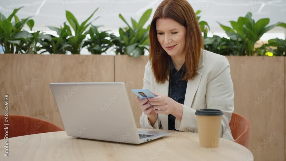 Smiling woman texting message on smartphone while sitting in cafe, communicating online, opened laptop in front of her, carton coffee cup standing beside. Concept of technology