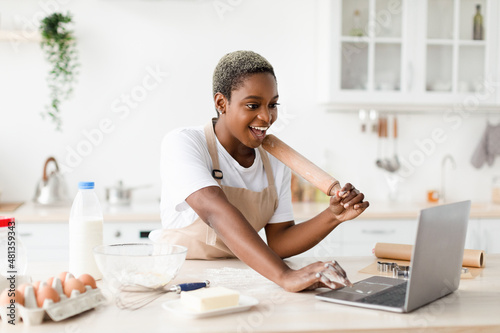 Smiling millennial cute black lady in apron prepare dough for baking pie or pizza and looks at laptop
