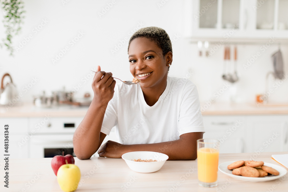 Smiling young attractive black woman eating porridge sits at table with cookies, juice and apples Smiling young attractive black woman eating porridge sits at table with cookies, juice and apples