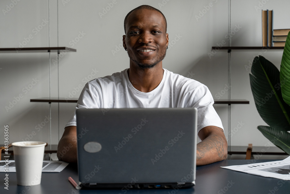Portrait of a smiling black man using a laptop and looking at the camera while enjoying work in an office interior Portrait of a smiling black man using a laptop and looking at the camera while enjoying work in an office interior