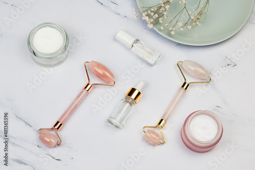 Face Massage pink quartz roller, face creams, face serum oil and flowers on white marble background. Skin care facial massage and relaxation. Anti-aging and lifting concept at home.