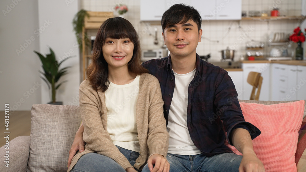 portrait of a smiling asian newly wed couple sitting close on sofa and looking at the camera in the living room at home portrait of a smiling asian newly wed couple sitting close on sofa and looking at the camera in the living room at home