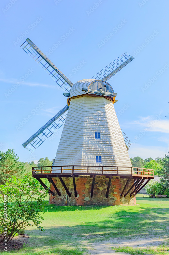 An old european wooden windmill on stone and brick basement, blue sky, green trees and grass
