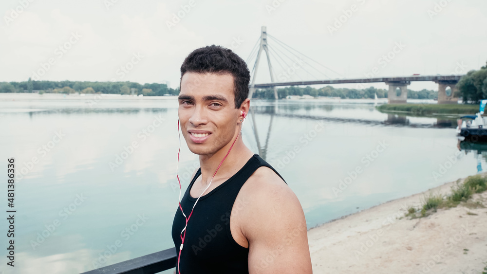 sportive bi-racial man in earphones smiling at camera near river.
