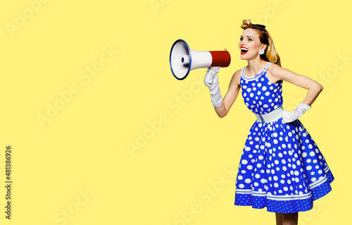 Blond haired woman holding red megaphone, shout advertise something. Girl in blue pin up style dress with mega phone loudspeaker. Yellow color background. Female model in retro fashion vintage photo.