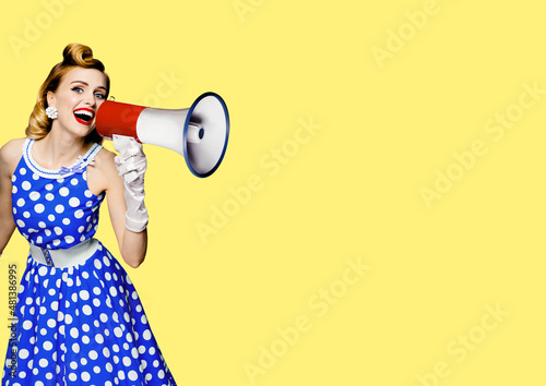 Portrait image of haired woman holding mega phone, shout advertising something. Girl in blue pin up style dress with mega phone loudspeaker. on yellow background. Beauty model in retro fashion concept