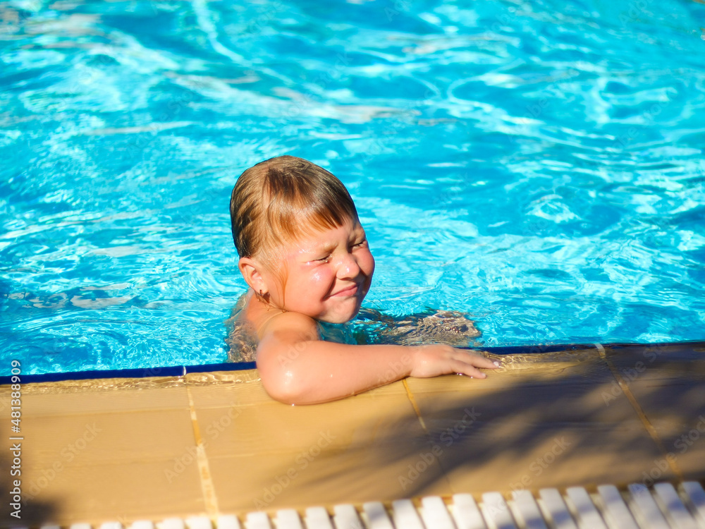 A cute little girl squints against the bright sun leaning on the side of the pool. Happy child swims and bathes in the pool on a sunny day. Selective focus.
