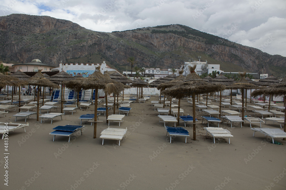 evocative image of a sandy beach with empty sunbeds and straw umbrellas 
with mountains and clouds in the background