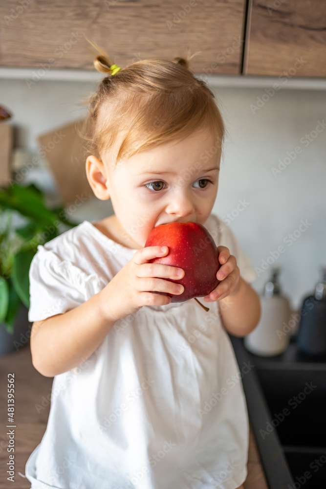 Baby girl blonde eating an apple in the kitchen, concept of healthy food for children